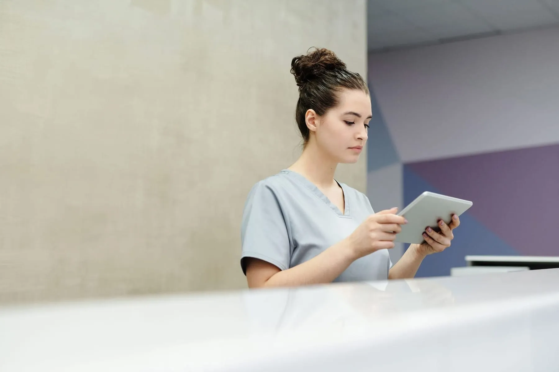 A receptionist in grey scrubs using a tablet at a clinic front desk